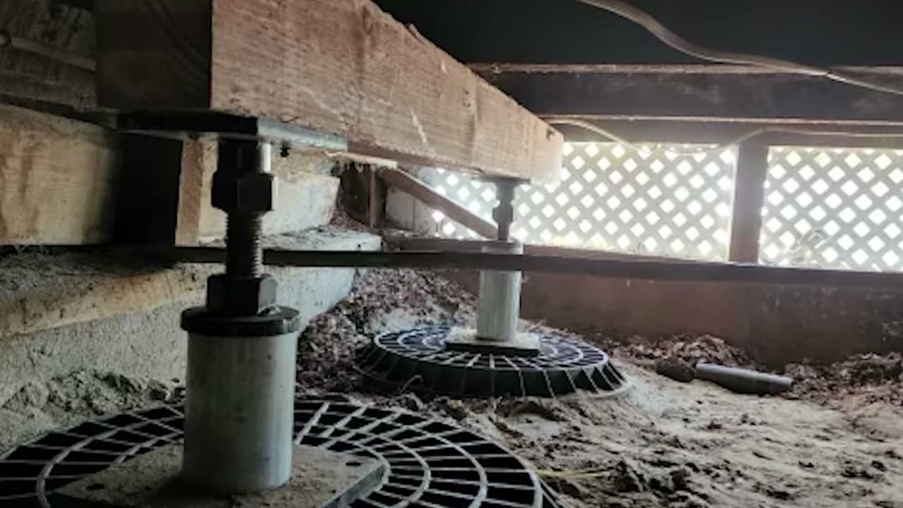Steel adjustable jacks on round footing pads holding up a wood floor beam in a crawl space