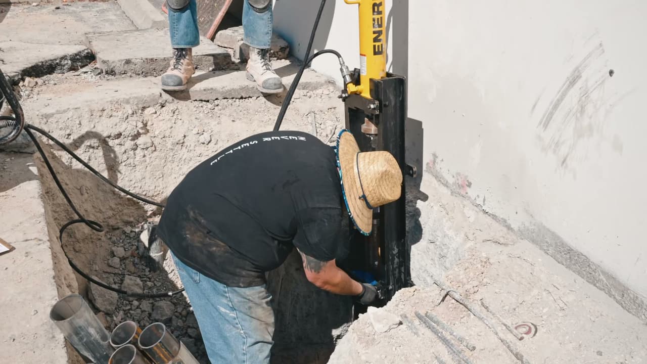 Crew member in a straw hat setting a hydraulic push pier into an excavation at the base of a white building wall in Mesa, Arizona