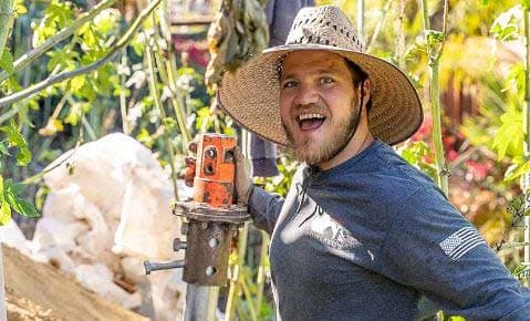 Smiling crew member in a wide brim straw hat holding a helical pier drive head in a garden