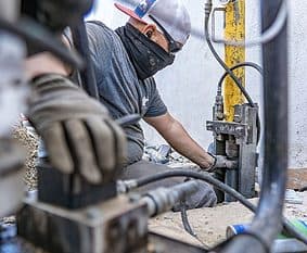 Crew member in a face covering working hydraulic pier driving equipment against the wall of a home