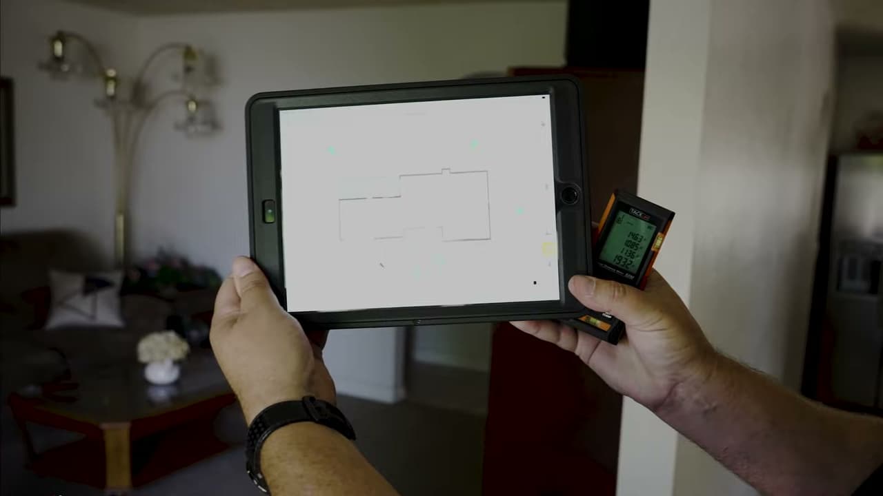 Hands holding a tablet with a floor plan sketch and a laser distance meter inside a living room