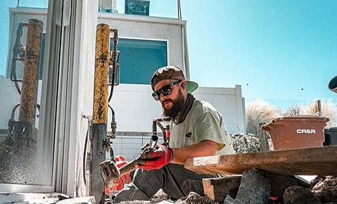 Crew member breaking up concrete with a jackhammer beside a sliding glass door, with hydraulic pier equipment standing against the wall