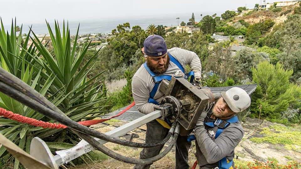Two crew members in safety harnesses running a hydraulic drive head on a steep hillside above the ocean