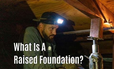 Crew member with a headlamp checking a steel support jack under a floor beam in a crawl space, titled What Is A Raised Foundation