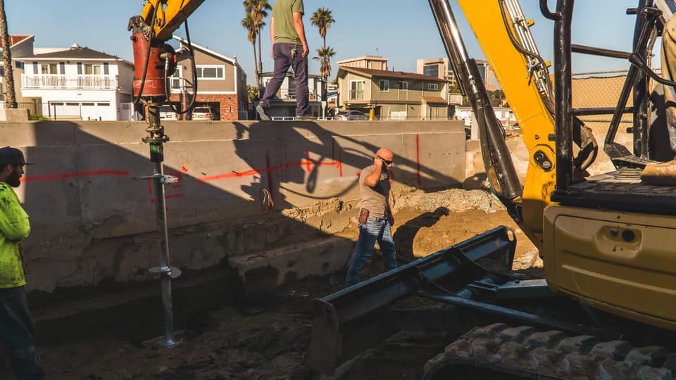 Crew driving a helical pier into the ground beside a concrete foundation wall, with an excavator working alongside