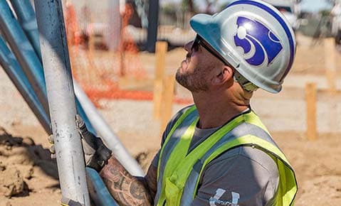 Worker in a hard hat and safety vest guiding a steel pile into place on a commercial site