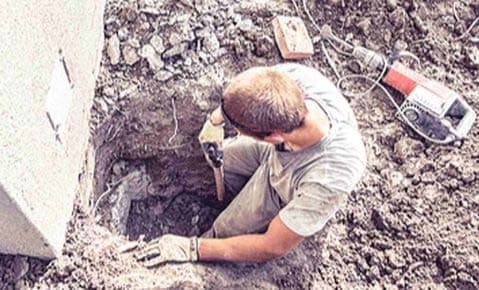 Worker digging an excavation pit beside a concrete footing, with a power tool at the edge