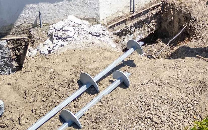 Two galvanized helical piers with screw shaped plates lying beside an open pit at a home's foundation