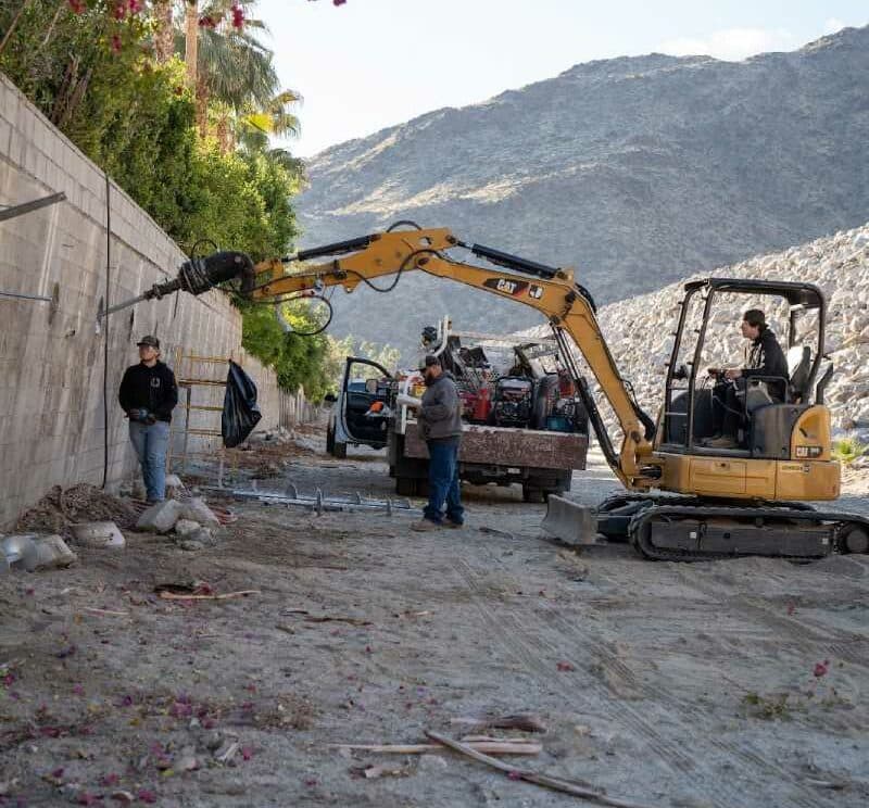 Dalinghaus Foundation Repair crew installing helical pier anchor systems into a concrete retaining wall with a mini-excavator