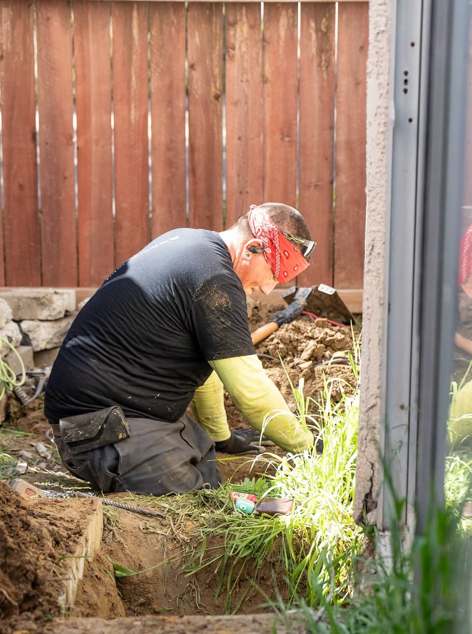 Crew member kneeling in a pier hole beside the stucco wall, reaching into the loose soil