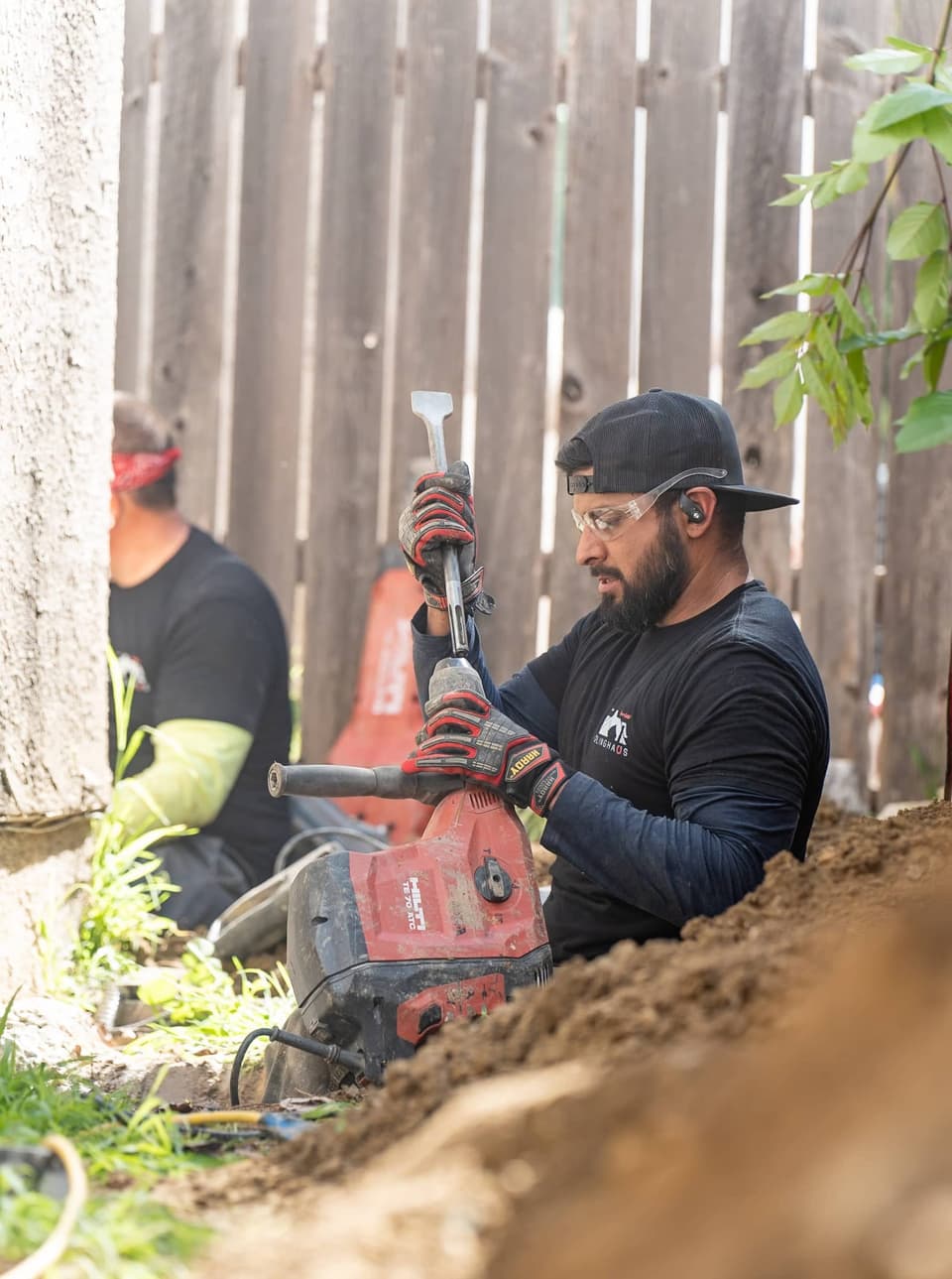 Worker in safety glasses fitting a chisel bit into a red demolition hammer beside the wooden fence