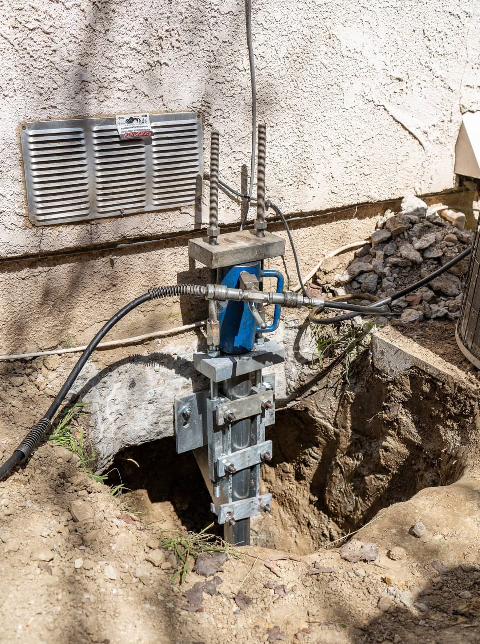 Blue hydraulic ram mounted on a push pier bracket below a foundation vent, hoses running to it
