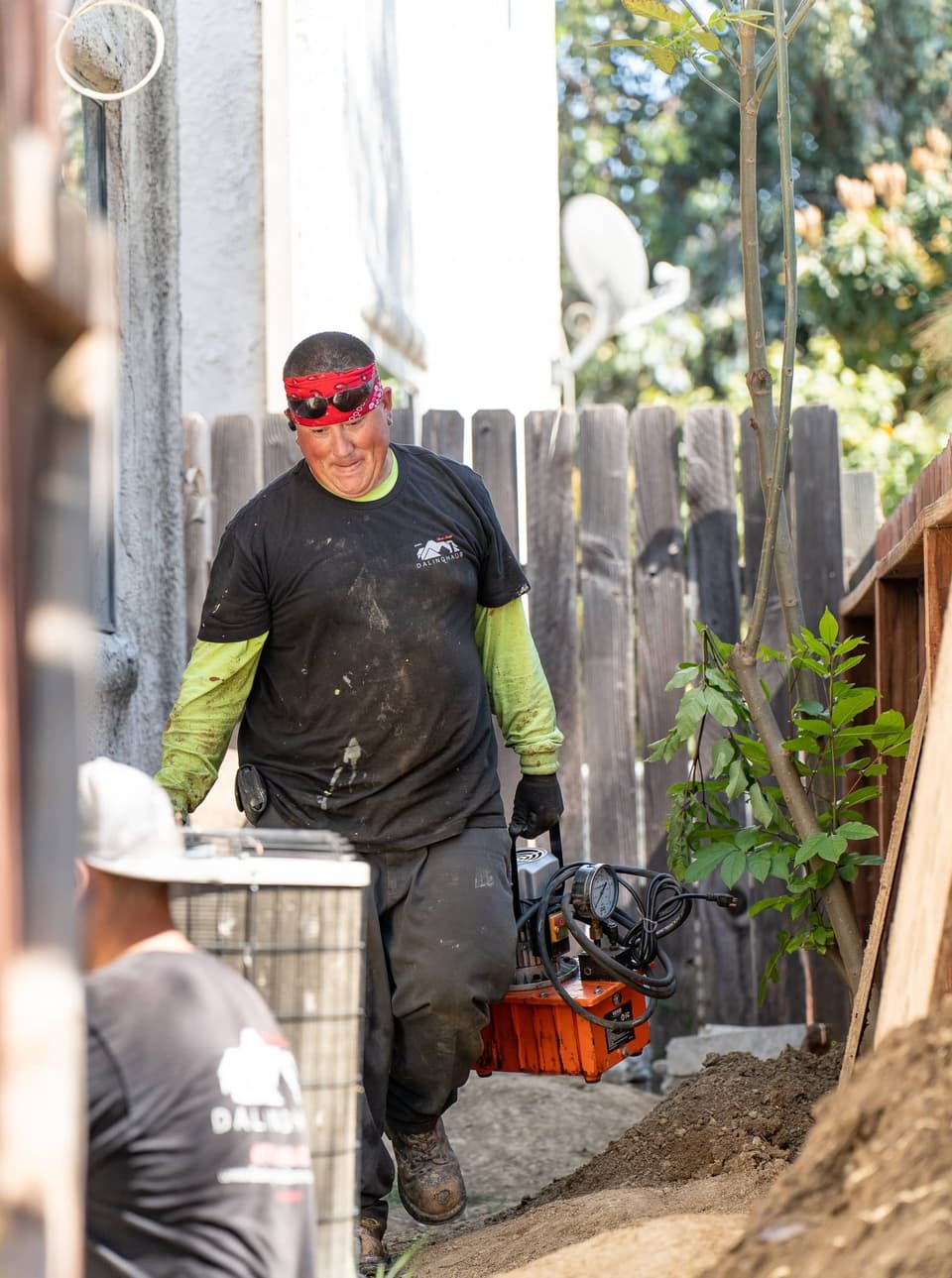 Two crew members in a narrow side yard, one carrying an orange hydraulic pump past the fence