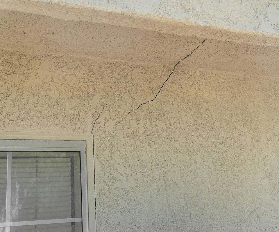 Diagonal crack in tan stucco running from a window corner up toward the eave