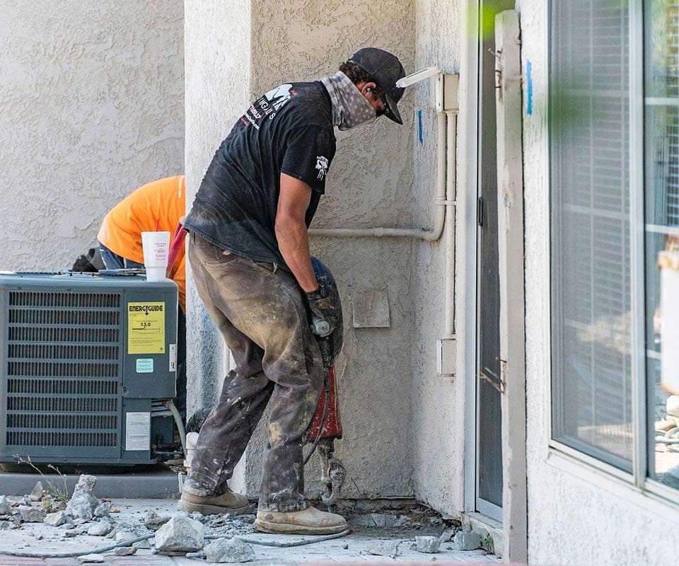 Crew member driving a demolition hammer into concrete at the base of a stucco wall beside an air conditioner