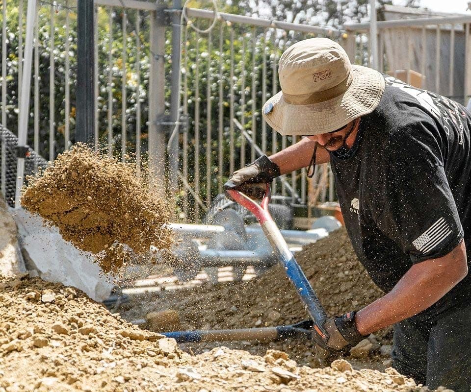 Crew member in a bucket hat and gloves flinging a shovel load of soil from a footing pit