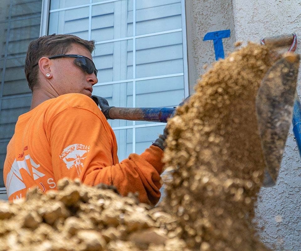 Crew member in an orange shirt and safety glasses shovelling soil below a window, numbered blue tape beside it