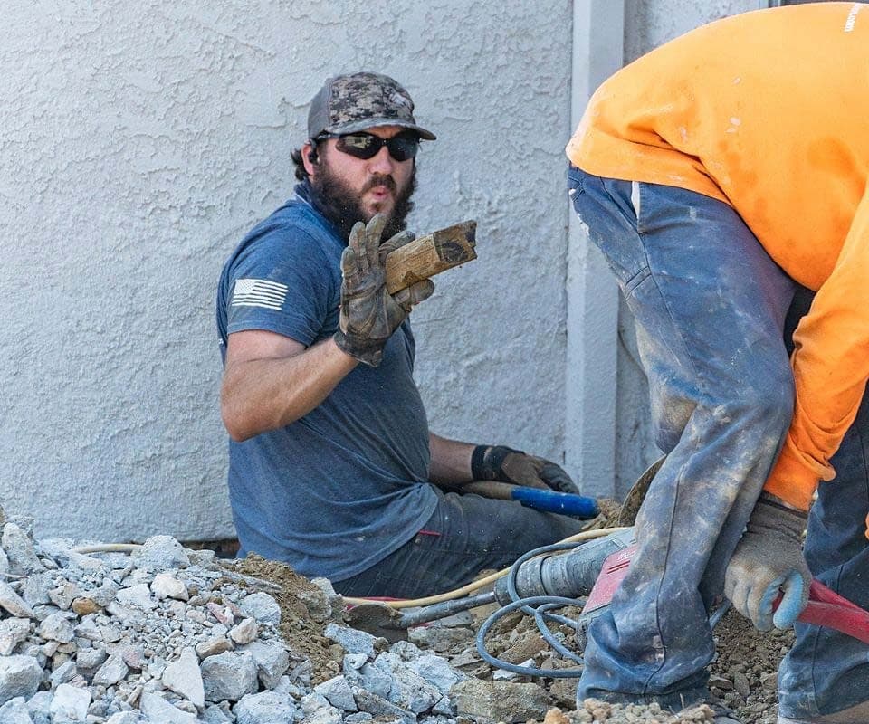 Bearded crew member seated in a footing pit holding up a chunk of soil, a second worker beside him