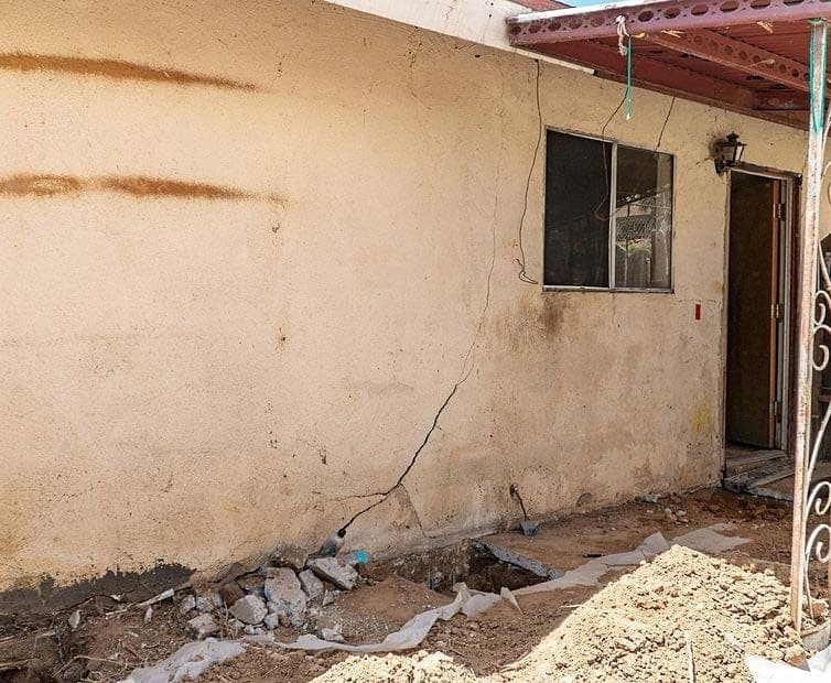 Long diagonal crack in tan stucco below a window, with a trench dug along the footing