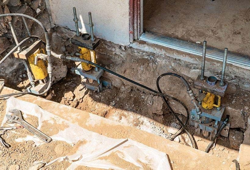 Two push pier brackets with hydraulic rams and hoses in a trench beside a door threshold