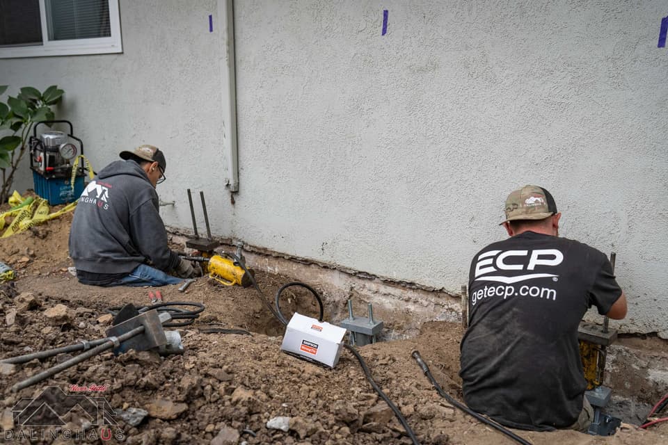 Two workers at helical pier brackets beside a house wall, a hydraulic pump and gauge on the ground behind them