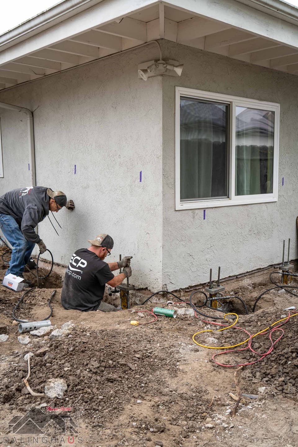 Two crew members working at a house corner where pier brackets and lift rams sit in shallow footing pits