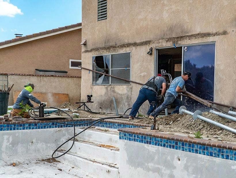 Three workers feeding a long steel helical shaft in through an open sliding door beside a drained pool