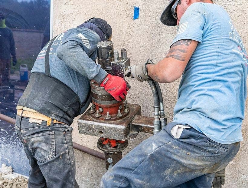 Two workers steadying a hydraulic torque motor on a helical pier shaft against a stucco wall