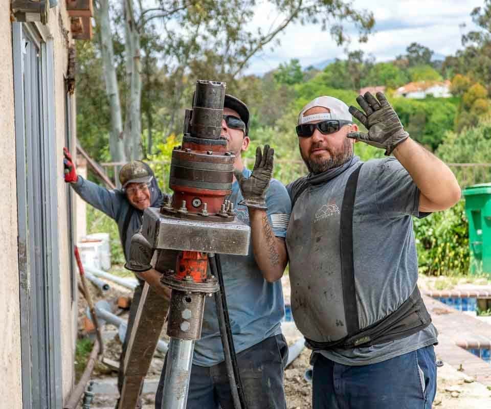 Two crew members wave at the camera beside a hydraulic helical drive head, a third behind them, hillside homes beyond