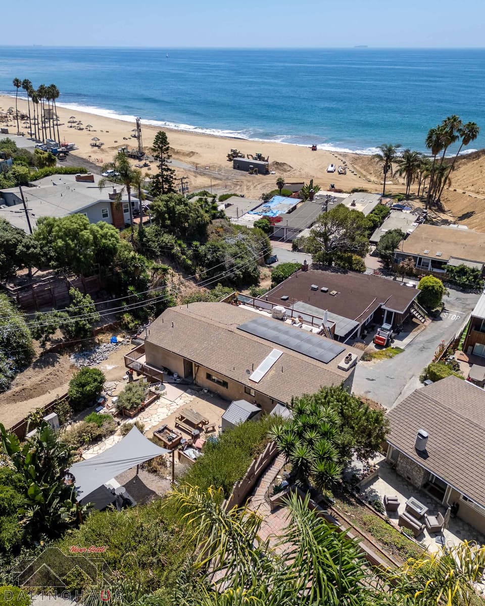 Drone shot over the home’s shingle roof and solar panels toward the beach, where a drill rig is working