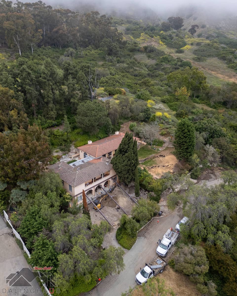 Wide foggy hillside aerial with the home small among trees and two work trucks parked on the road below