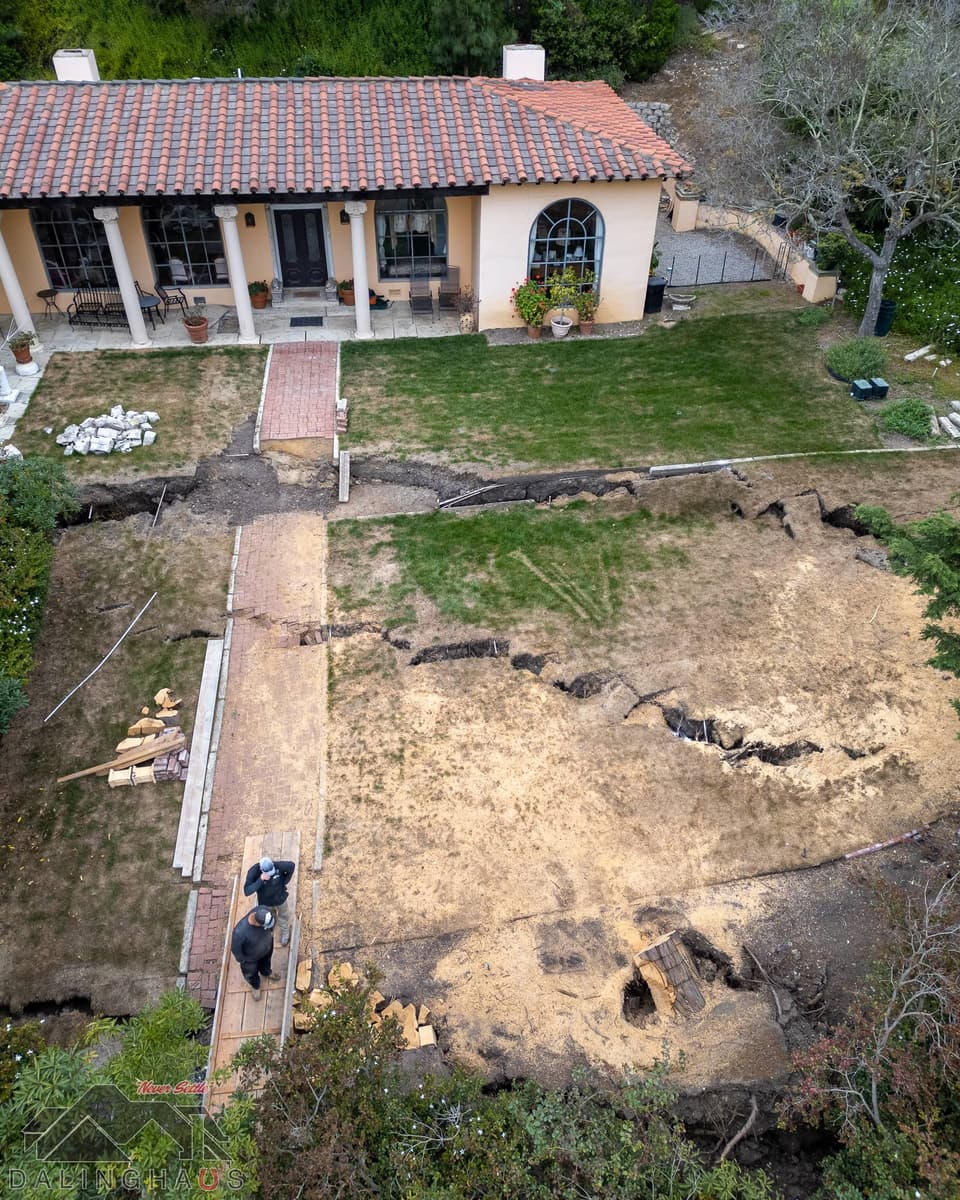 Two crew members on a plywood walkway beside a broken brick path and a split lawn