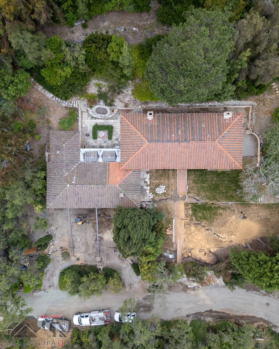 Straight down drone view of both wings of the home, steel beams and an excavator at left, cracked yard at right