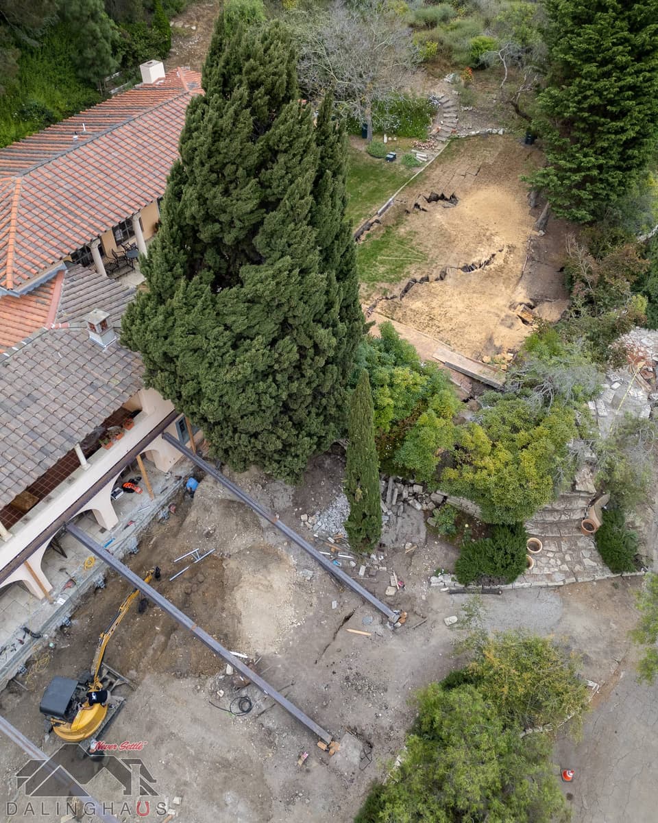 Overhead of steel beams under the home, an excavator at work and a cracked lawn beyond the cypress trees