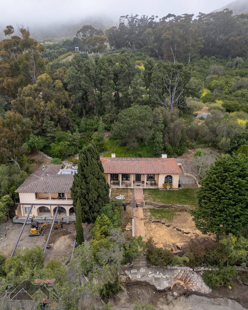 Aerial of the home from the hillside side, beams and excavator under the far wing, workers in the yard