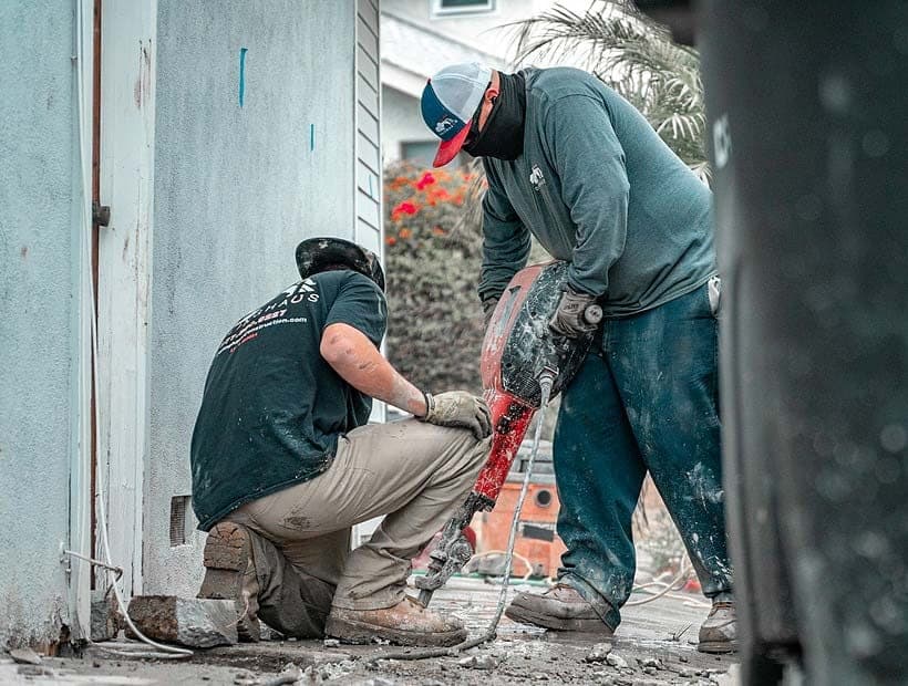 Two crew members breaking out concrete with a jackhammer at the base of a stucco wall