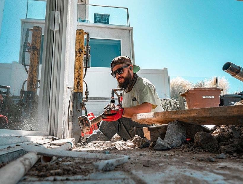 Crew member in sunglasses and a cap holding an impact wrench at ground level below two yellow hydraulic rams