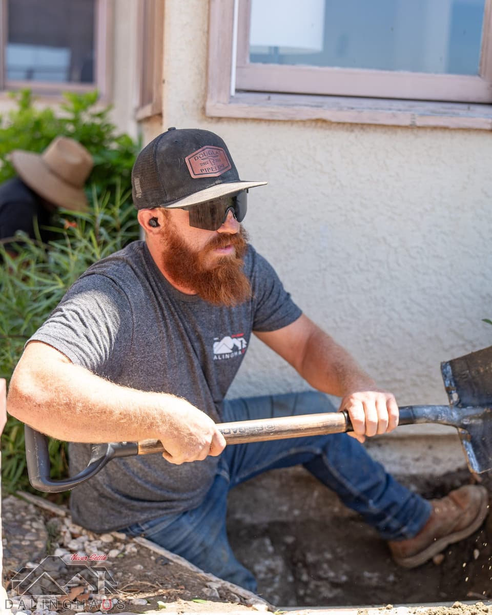 Crew member in a ball cap and sunglasses shoveling a hand dug pit at the footing beneath a window