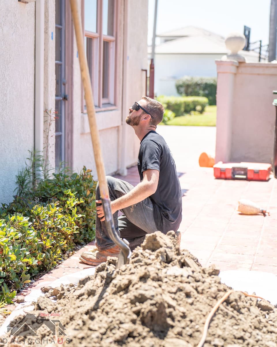 Crew member crouched by a pile of excavated soil on plastic sheeting along the paver patio, shovel propped beside him