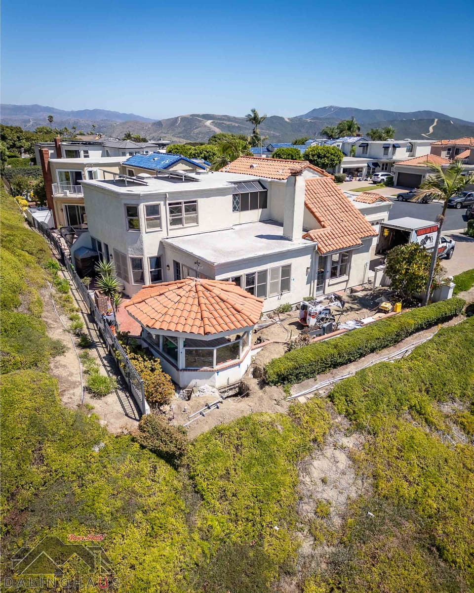 Angled drone shot of the rear yard with a track loader, tools and open excavation beside an octagonal sunroom