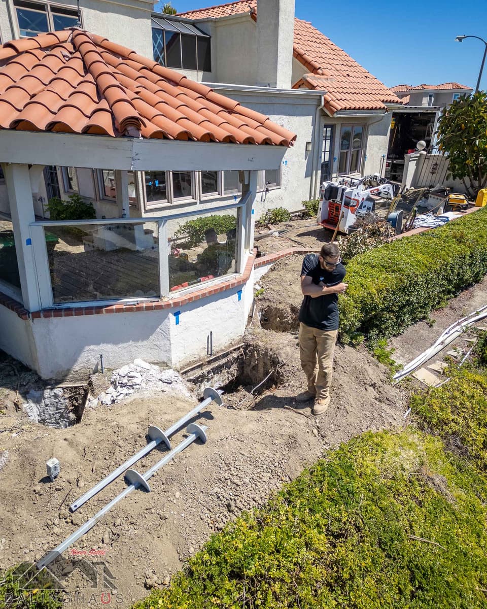 Crew member standing beside open pits at the sunroom footing with three helical pier sections laid on the dirt