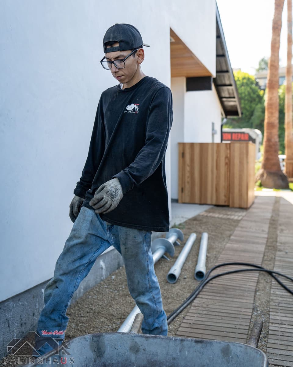 Crew member walking down a gravel side path past helical pier sections laid beside a white home