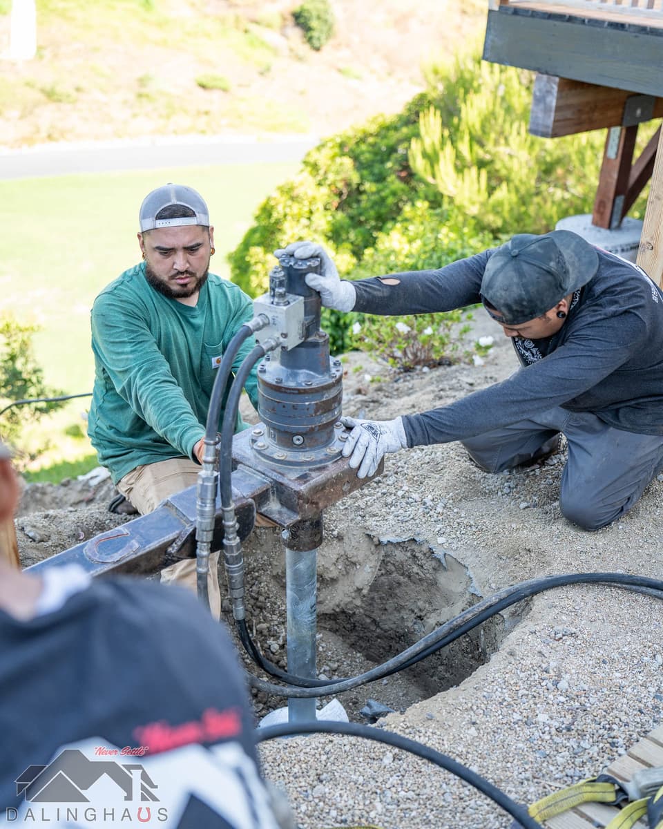 Two crew guiding a hydraulic drive head as a helical pier enters a hand dug pit beside the deck