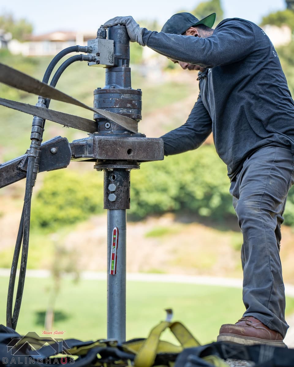 Crew member steadying a hydraulic drive head with a level taped to the pier shaft, green slope behind