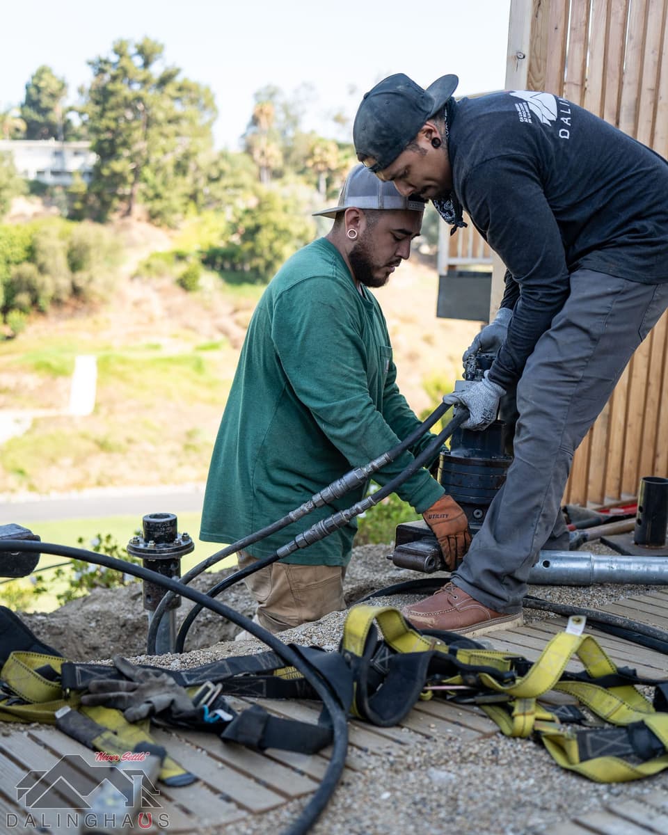 Two crew connecting hydraulic hoses to a drive head with a pier stub in the ground and hillside behind