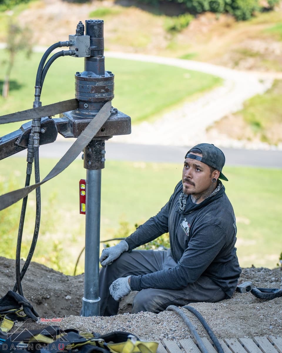Crew member seated beside a helical pier under a drive head, level on the shaft, golf course path below