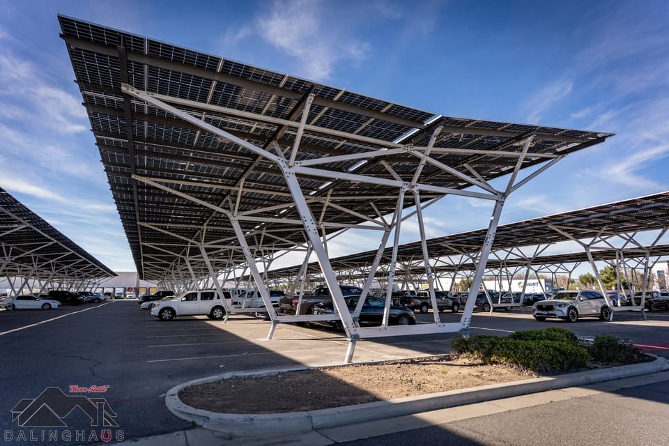 Finished solar canopy over a full parking lot, white branching steel columns fanning out beneath a continuous deck of solar panels, further canopy rows receding on both sides under a blue sky.