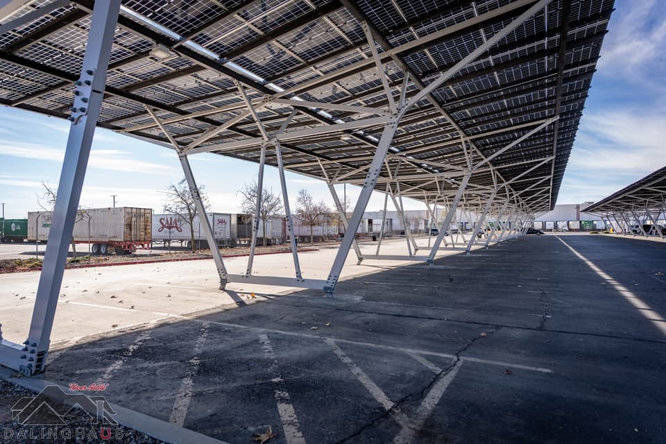 View from beneath the finished solar canopy looking down an empty parking aisle, white steel columns and the underside of the panel deck overhead, semi trailers and a shipping container along the back fence.