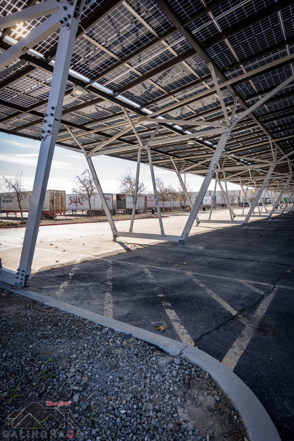 Low view under the finished canopy with the panel deck overhead and white steel columns receding, semi trailers and a shipping container visible past the edge of the structure.