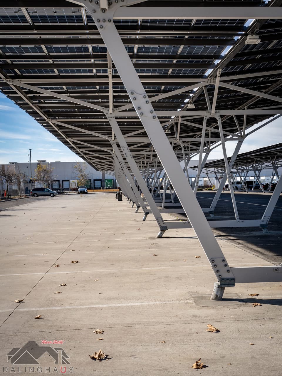 Perspective down a long row of white steel canopy columns under the solar panel deck, loading docks and parked vehicles at the left, empty concrete with scattered dry leaves in the foreground.